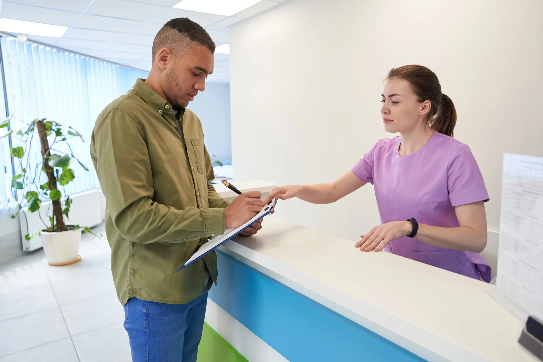 African American man standing by reception counter and writing on clipboard while woman hospital worker pointing at papers